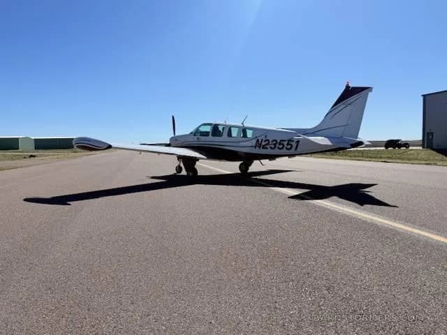 A white and red Beechcraft Bonanza aircraft is parked on an asphalt runway, viewed from the rear-left.
