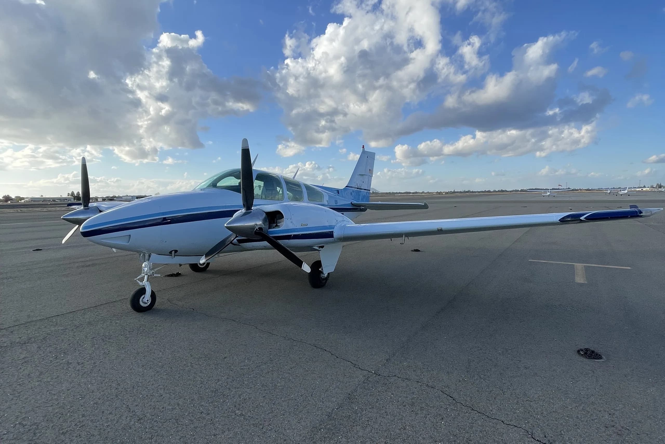 A white and blue twin-engine propeller aircraft is parked on an asphalt tarmac under a partly cloudy sky.