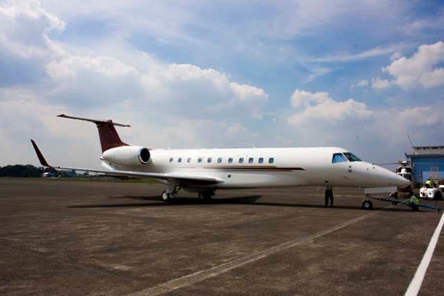 A white business jet with red and gold stripes on the tail and fuselage, parked on an airfield with ground equipment nearby.