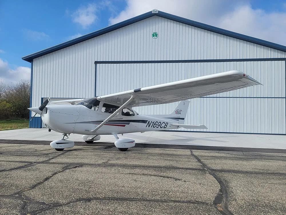 A white Cessna Skyhawk N169CB with black and red stripes is parked on an asphalt ramp in front of a white hangar under a partly cloudy sky.