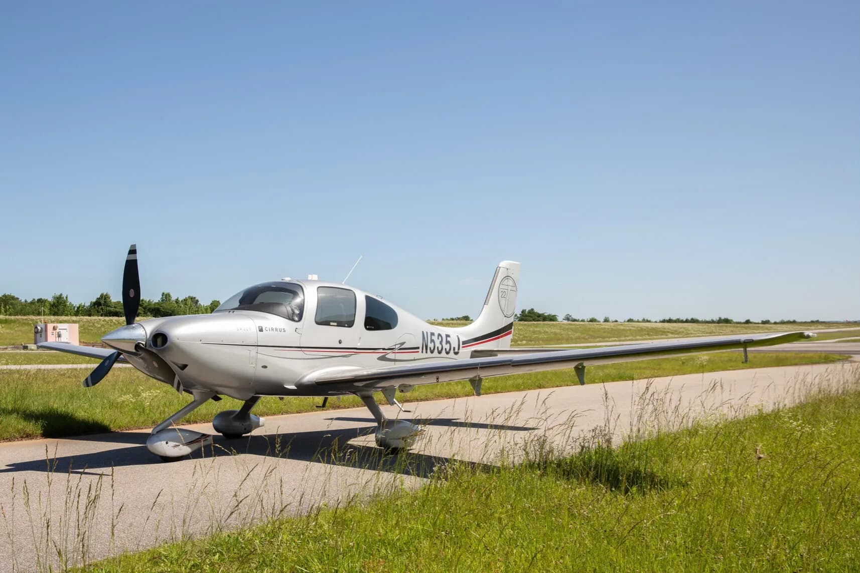 A silver Cirrus SR22T aircraft with registration N535J is parked on an asphalt strip next to green grass under a clear blue sky.