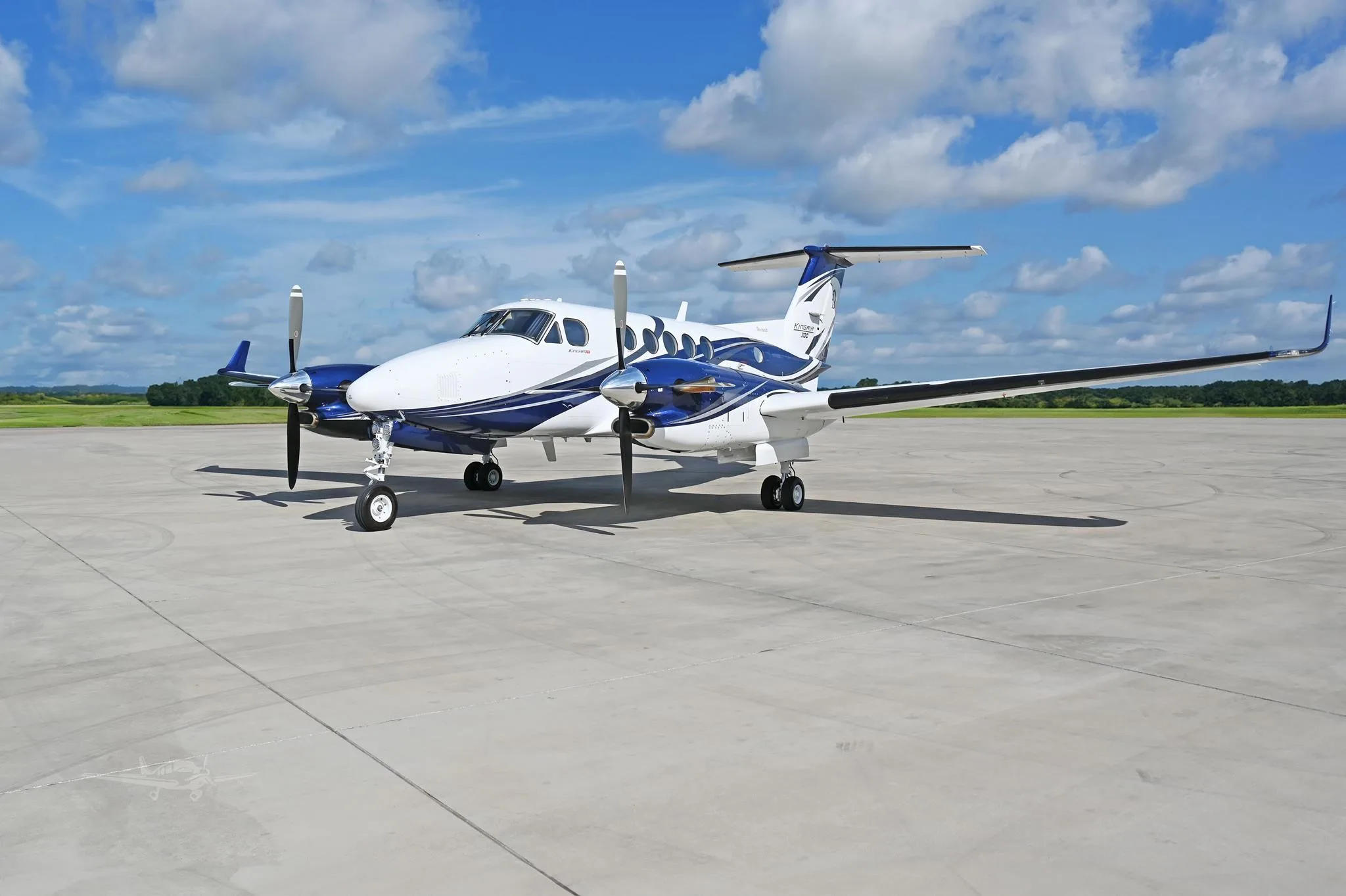 A white and blue Beechcraft King Air 350 twin-turboprop aircraft is parked on a concrete tarmac under a partly cloudy sky.