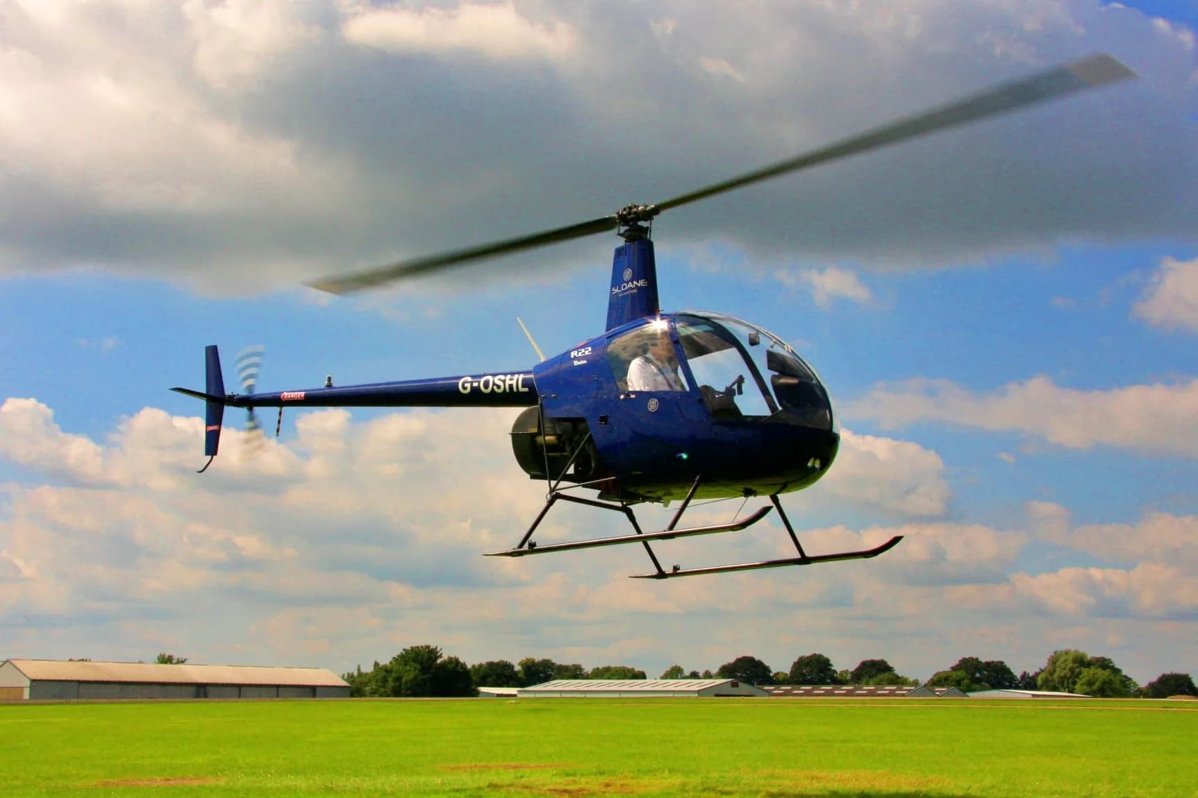 A blue Robinson R22 helicopter in flight against a cloudy sky.