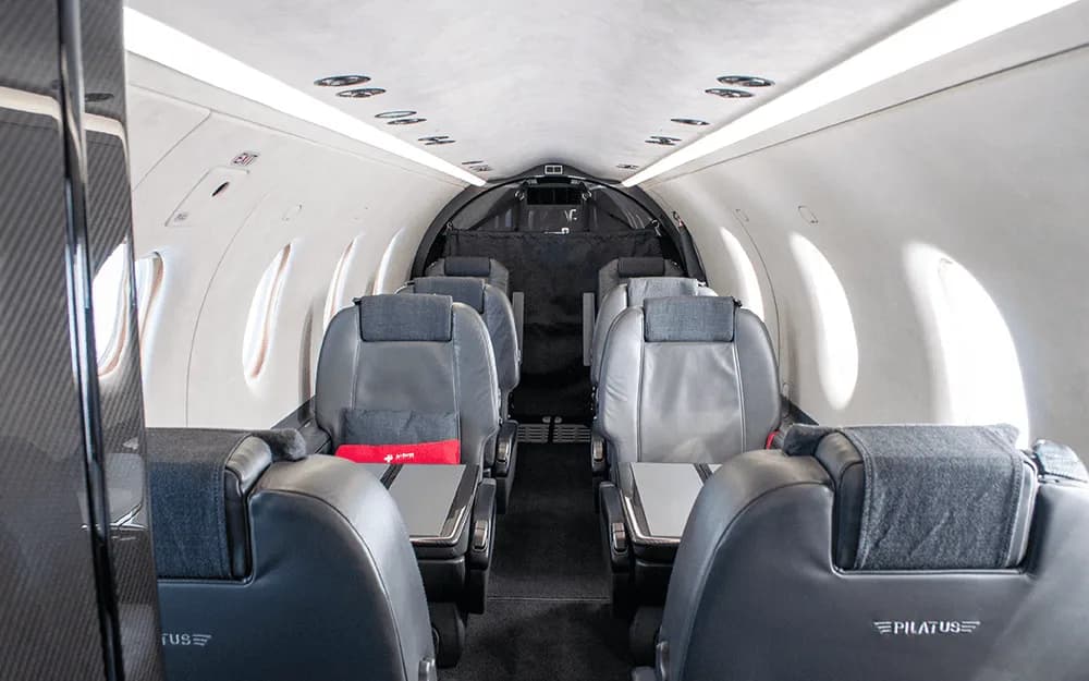 The spacious cabin interior of a Pilatus PC-12 aircraft, showing rows of dark grey leather seats with fold-out tables.