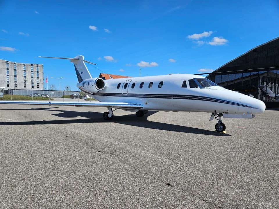 A white Cessna Citation business jet with a black stripe is parked on an asphalt tarmac under a blue sky, with buildings in the background.