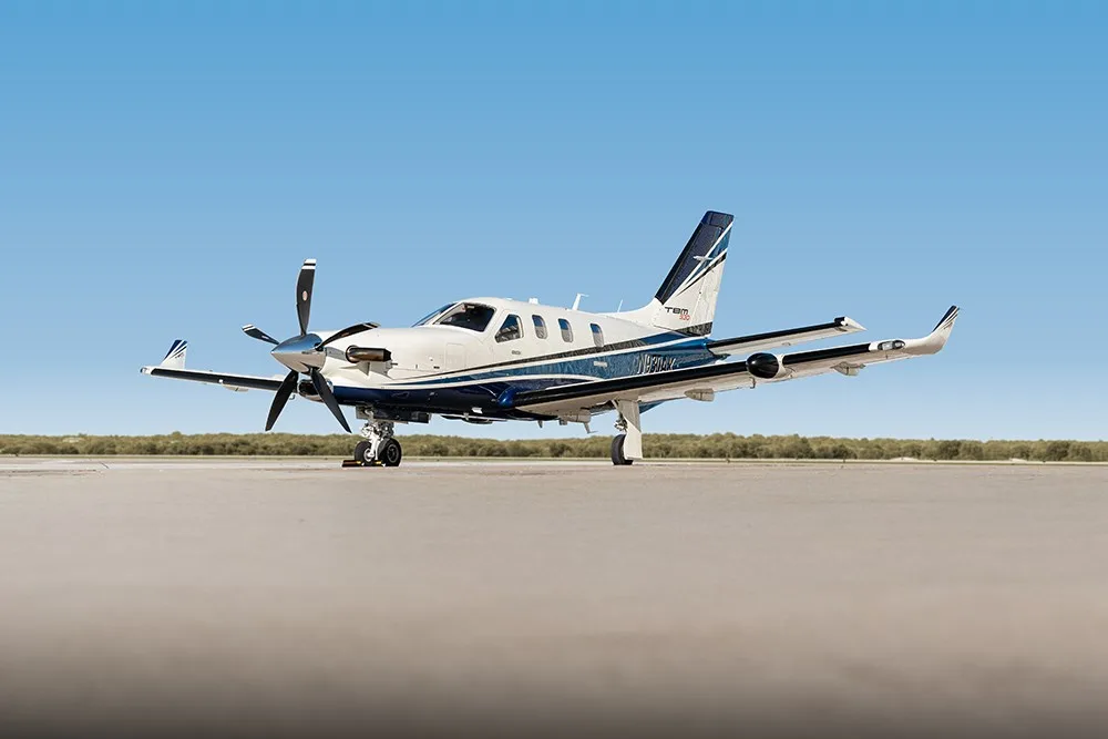A TBM 930 aircraft with registration N930AK in white and blue livery parked on an airfield, viewed from the front.