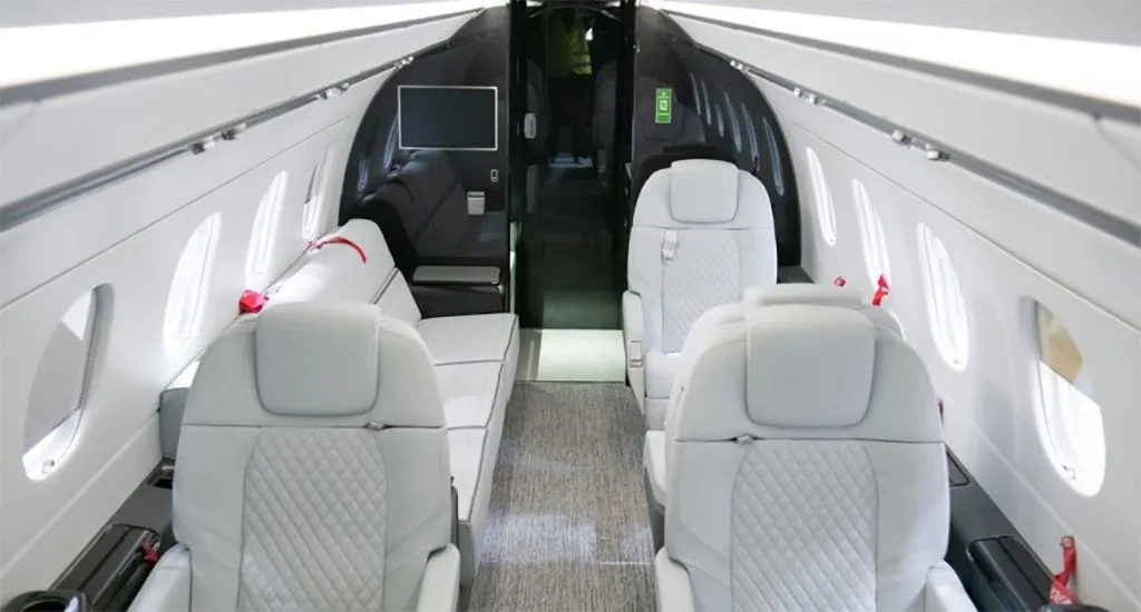 A spacious aircraft cabin interior with light grey seating, including individual seats and a side couch, looking towards the front.