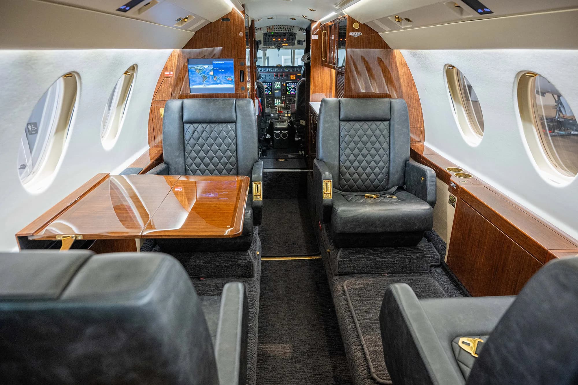 The cabin interior of a Dassault Falcon 900EX business jet showing a polished wooden table and dark quilted leather seats.
