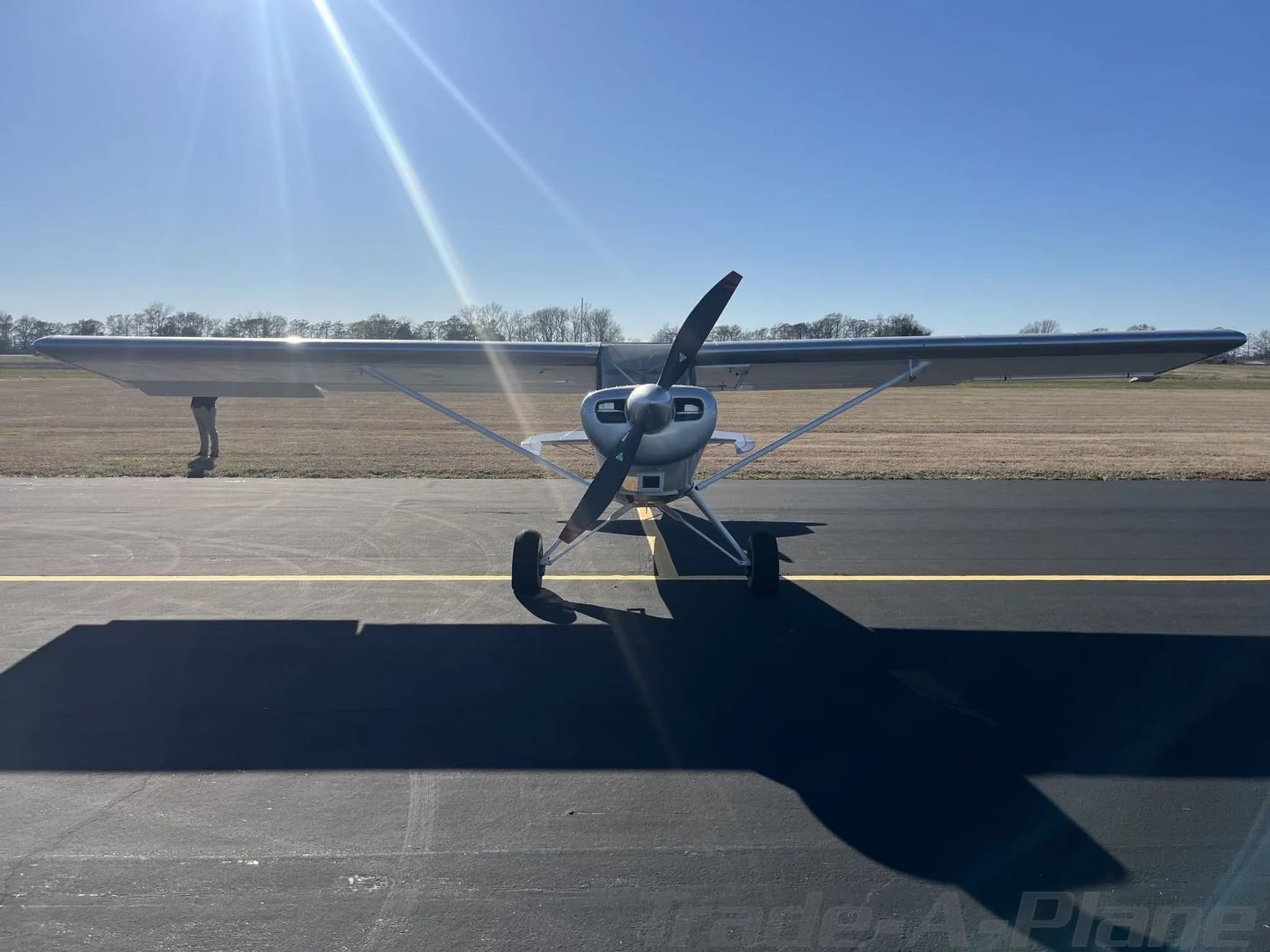 Front view of a silver Bearhawk Patrol aircraft with a propeller, parked on an asphalt tarmac under a bright sun.