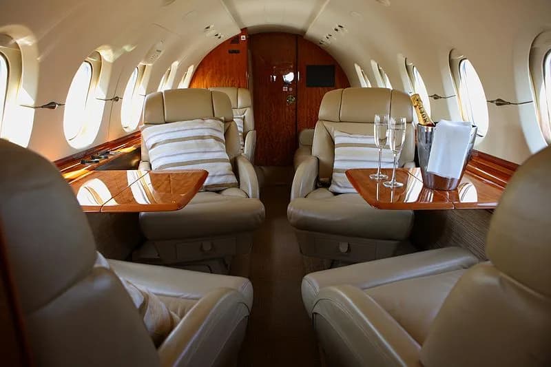 A luxurious aircraft interior with tan leather seats, wooden accents, and champagne flutes on a table.