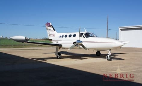 A white twin-engine propeller aircraft, N75SK, with purple and black stripes on its tail, parked on a tarmac next to a hangar under a clear blue sky.