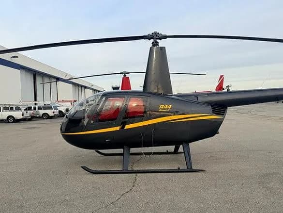 A black Robinson R44 Raven II helicopter with a yellow stripe, parked on an airfield next to a hangar.