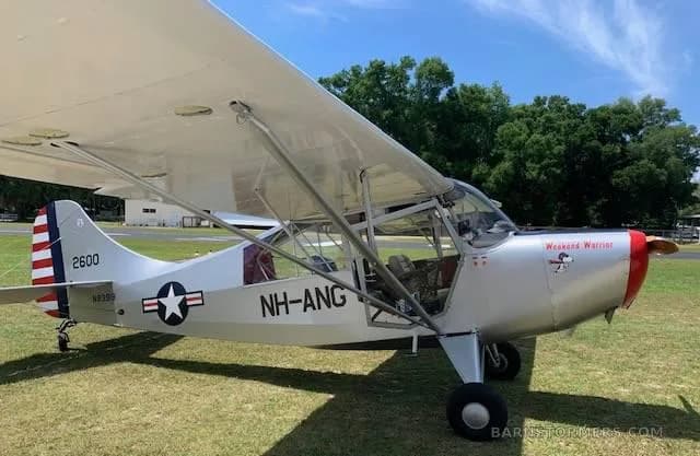 A silver Aeronca L-16 Liaison aircraft with "Weekend Warrior" nose art and US Air National Guard markings, parked on a grassy field.