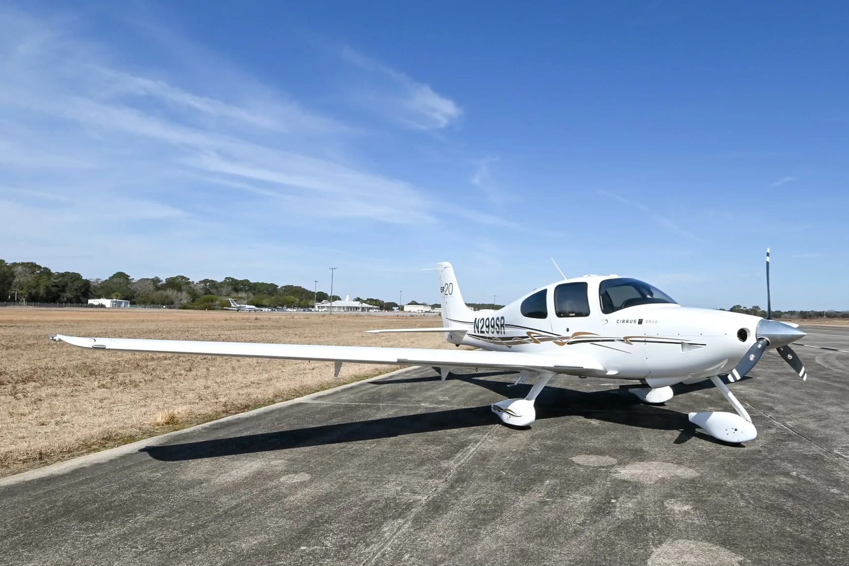 A white Cirrus SR20 aircraft with black and gold accent stripes, registration N299SR, parked on an airfield with buildings in the distance.