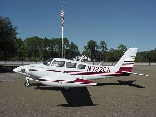 A Piper Apache aircraft with a white and maroon paint scheme is parked on the grass with an American flag in the background.