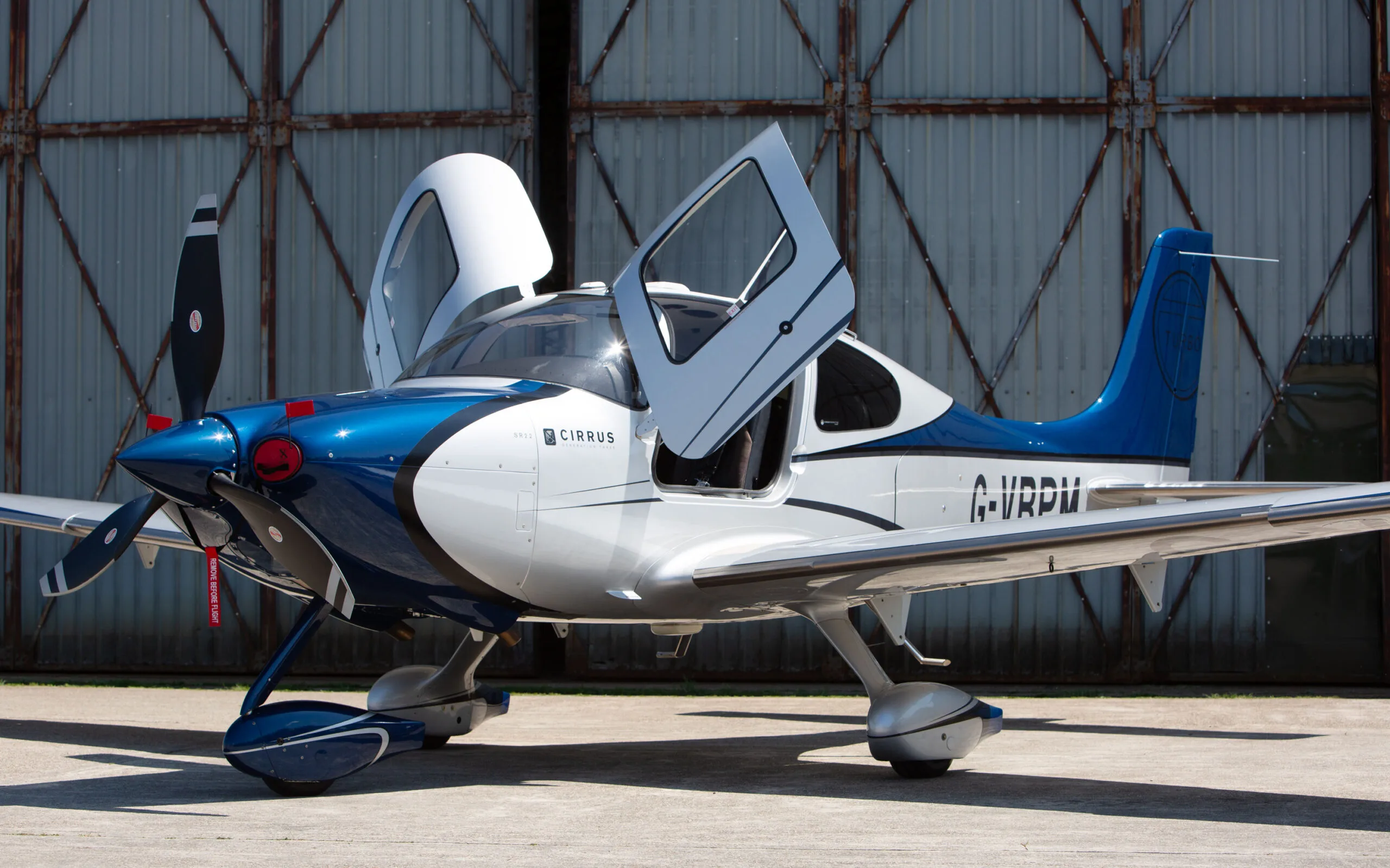 A blue and white Cirrus SR22 aircraft with registration G-VBPM and its gull-wing doors open, parked on an airfield apron.