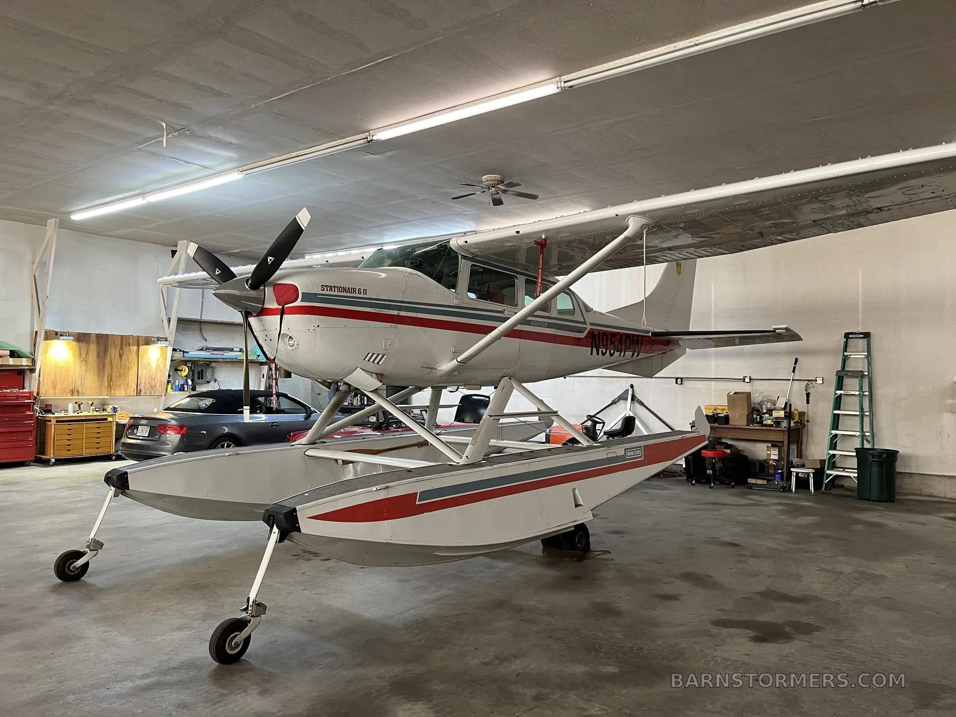 A white Cessna Stationair 6 II floatplane with red and blue stripes, registration N954PW, viewed from the front-left in a hangar.