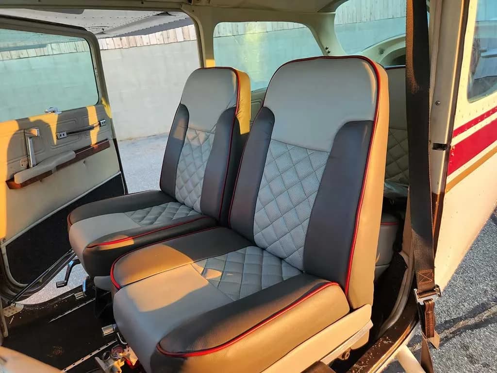 The front cabin seats of the aircraft, upholstered in gray and white with a quilted pattern and red piping, viewed through an open door.