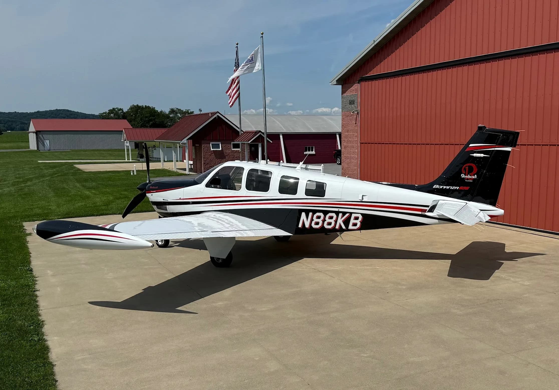 A white and black Beechcraft Bonanza with red stripes, tail number N88KB, parked on a concrete pad next to a red hangar.