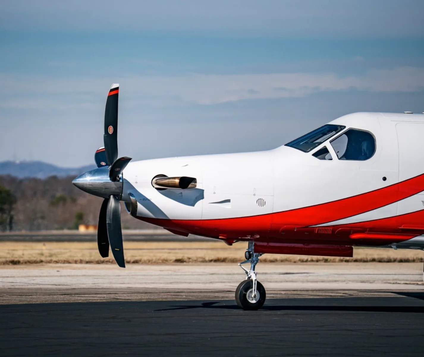 A close-up side view of the front section of a Pilatus PC-12 aircraft with a red and white paint scheme and a three-bladed propeller.
