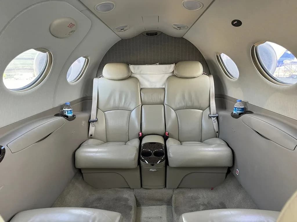 The interior cabin of a private jet showing two light brown leather seats with a small table between them and water bottles in cup holders.