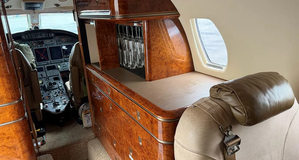 A view inside the business jet cabin showing a polished wood cabinet with glassware, with the cockpit visible in the background.
