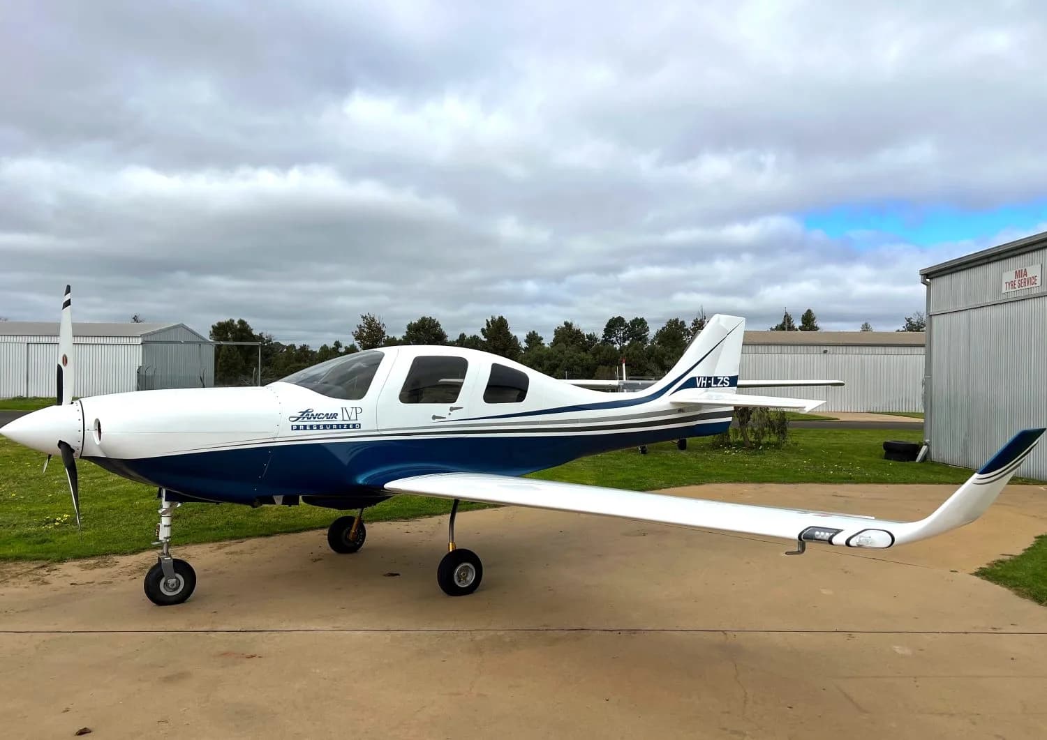 A white and blue Jabiru J430 aircraft (VH-LZS) with a propeller, parked on an asphalt surface next to a hangar under a cloudy sky.