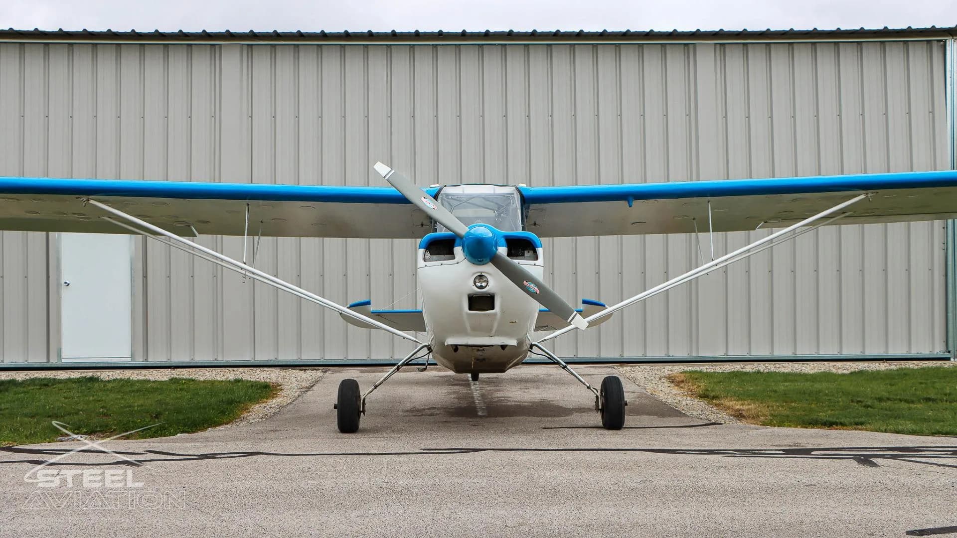 Head-on view of a white and blue Bellanca 7GCBC Citabria aircraft with a propeller, parked in front of a hangar.