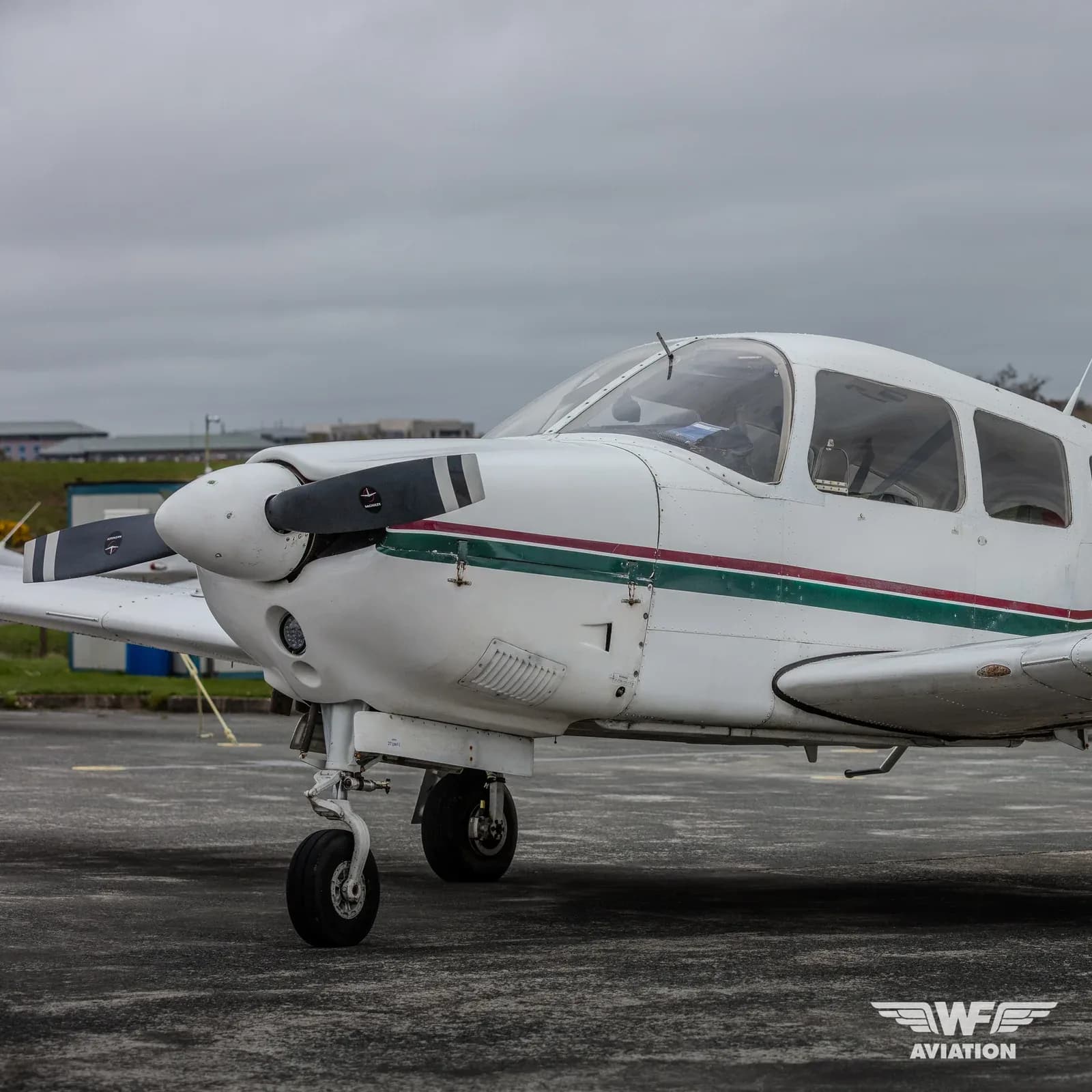 A white single-engine propeller aircraft with green and red stripes on the fuselage, parked on an asphalt tarmac under a cloudy sky.
