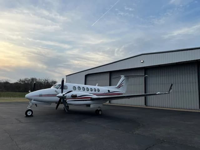 A white Beechcraft King Air with red and black stripes parked on an tarmac in front of a hangar, viewed from the left side.