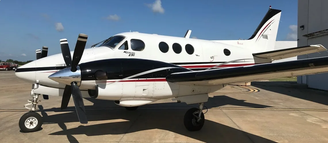 A white Beechcraft King Air E90 with black and red stripes is parked on an airport ramp under a clear blue sky.