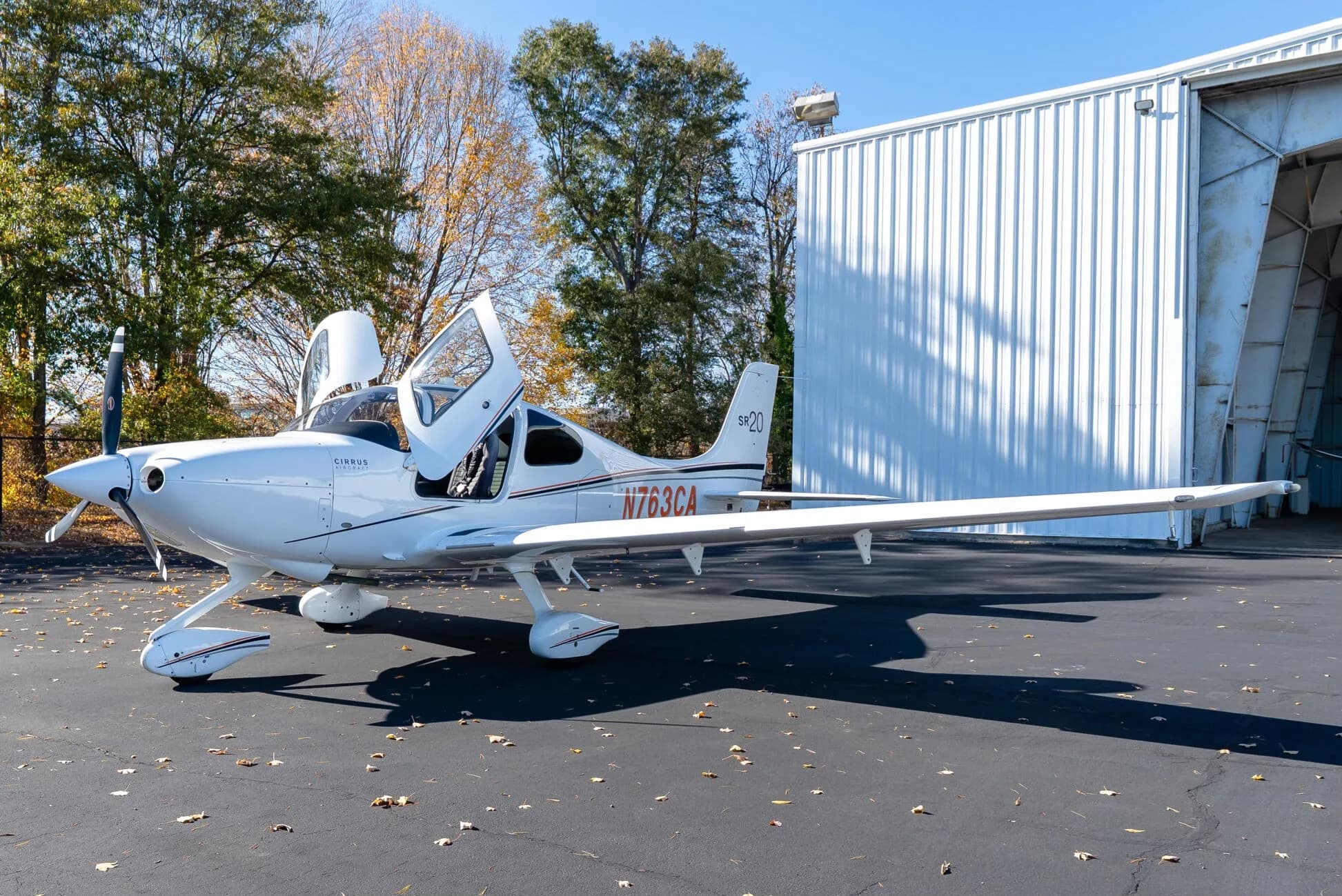 A white Cirrus SR20 aircraft with its doors open is parked on an asphalt tarmac next to a hangar, with autumn trees in the background.