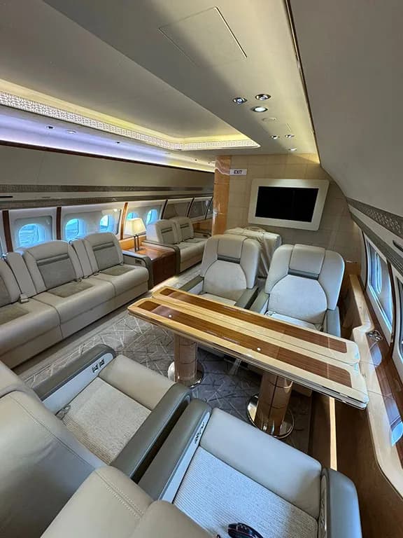 The spacious main cabin of a private jet includes multiple seating areas, a dining table, and a wall-mounted television.