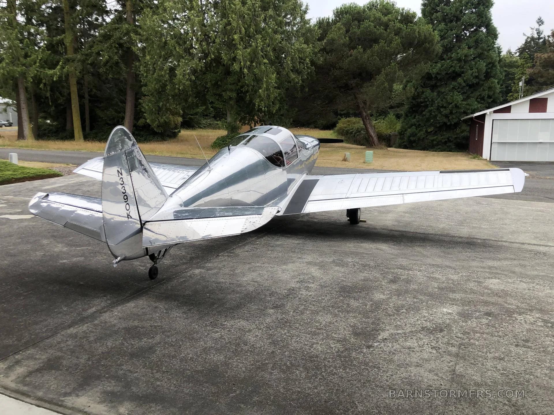 A shiny silver single-engine aircraft with tail number N3390K is parked on a concrete pad outside a hangar, surrounded by trees.