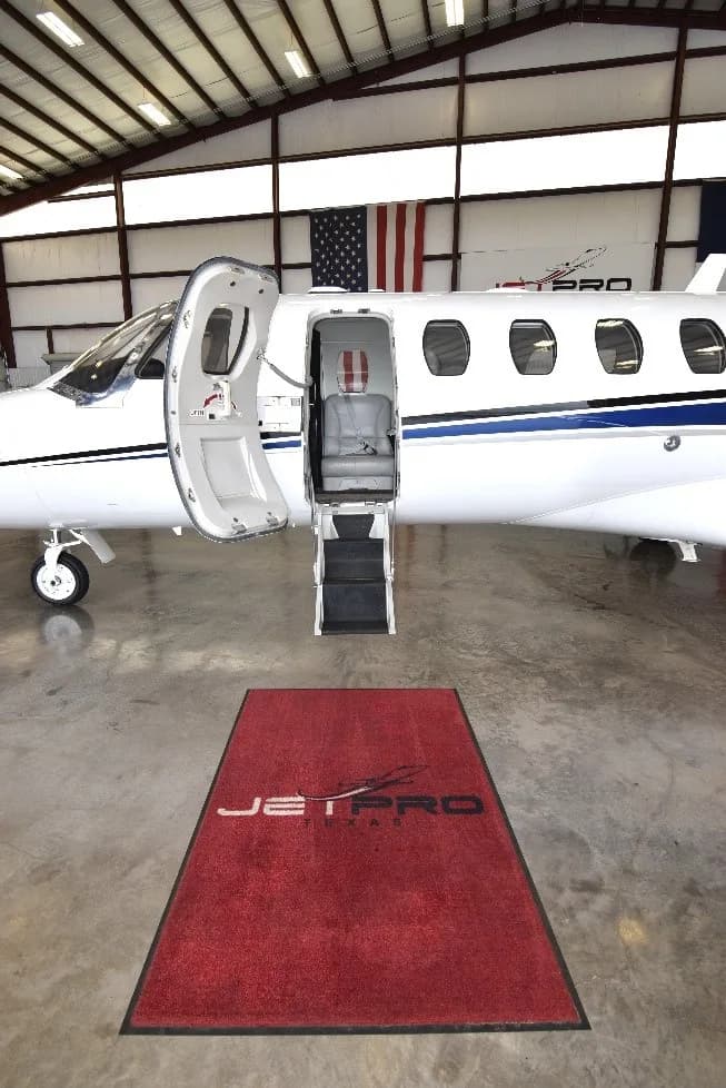 The open passenger door and airstairs of a white Cessna Citation CJ3 jet, with a "JET PRO Texas" mat on the hangar floor.