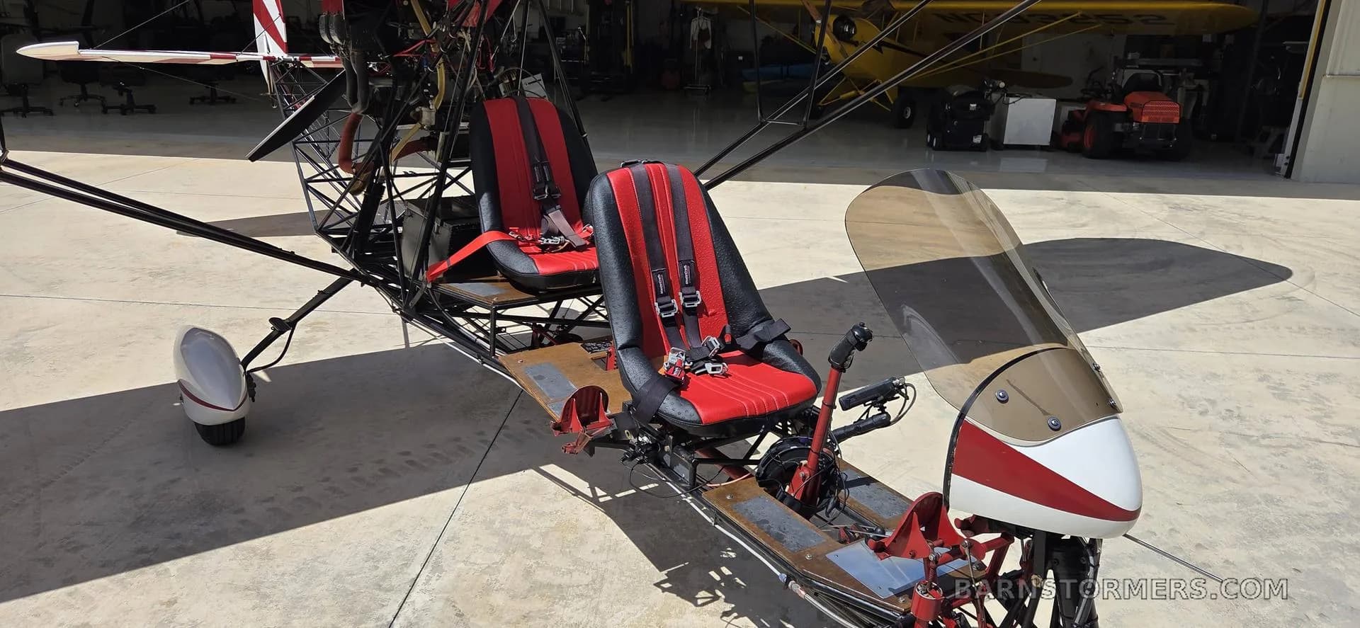 The red and black seats and front controls of an open-frame ultralight aircraft are visible, parked in a hangar.