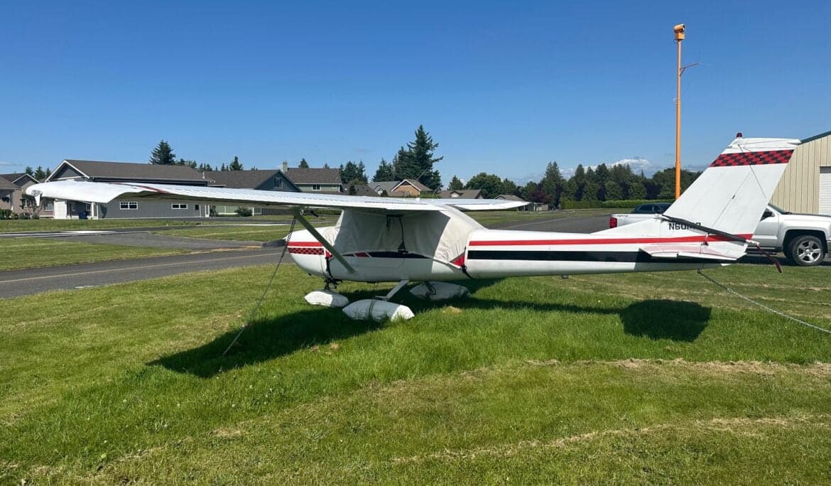 A Cessna 150 aircraft is parked on the grass with a white and black paint scheme and red accents.