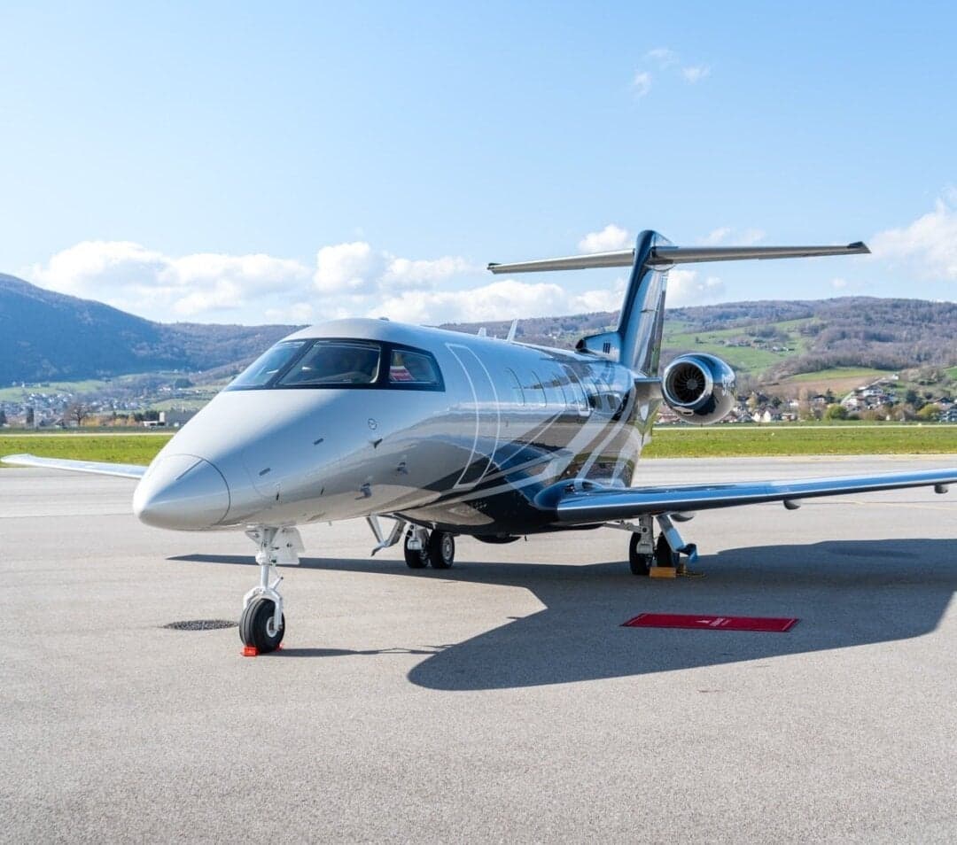 A silver and black Embraer Phenom 300 private jet parked on an airport tarmac with mountains in the background.