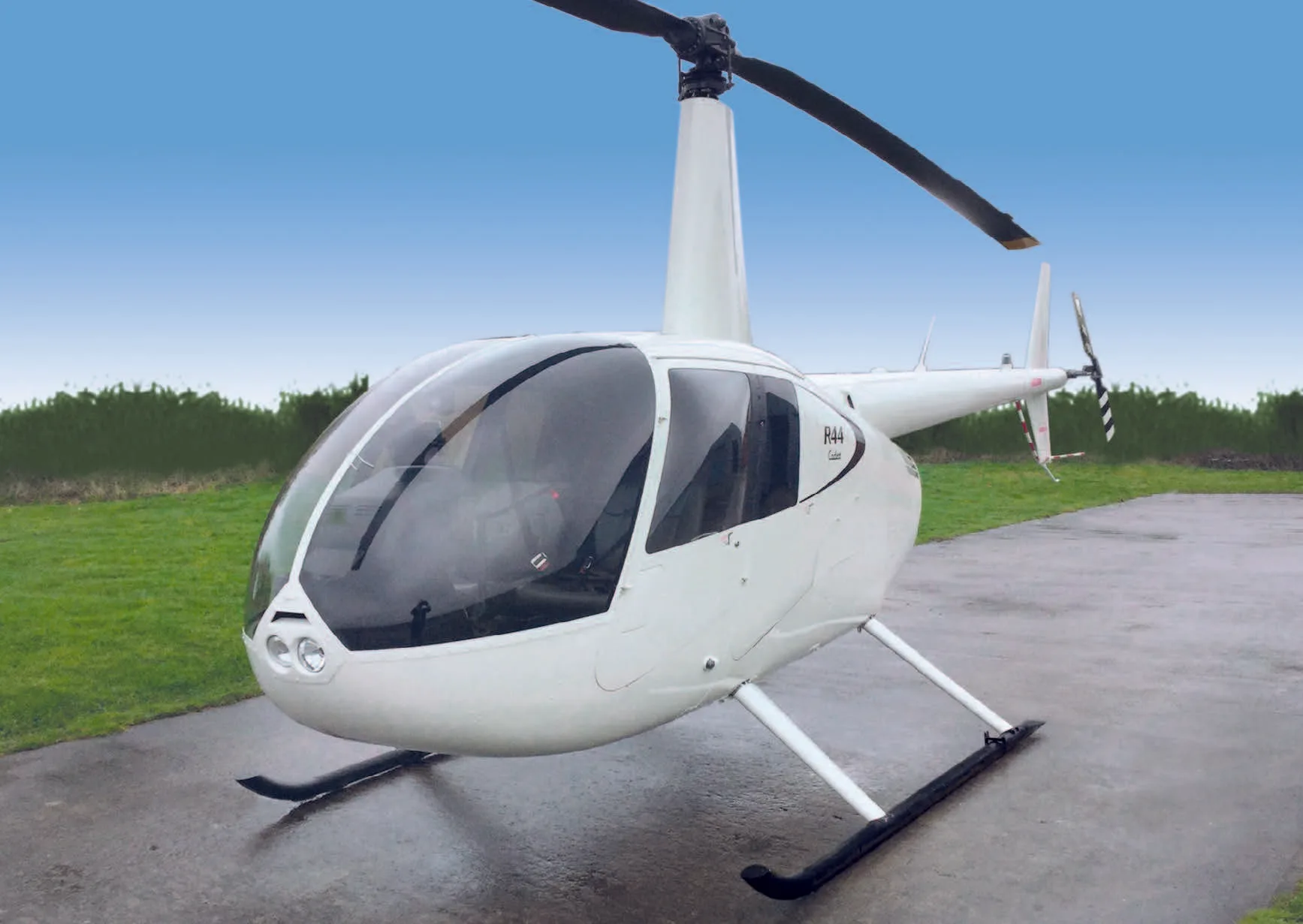 A white Robinson R44 helicopter with black skids sits on a tarmac with green grass and a blue sky in the background.