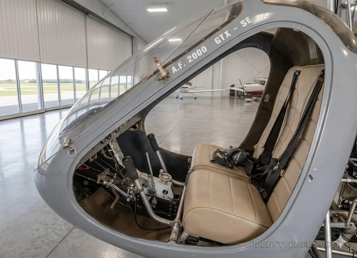 The interior of a grey RAF 2000 GTX-SE autogyro shows two beige seats and controls, with other aircraft visible outside a hangar.