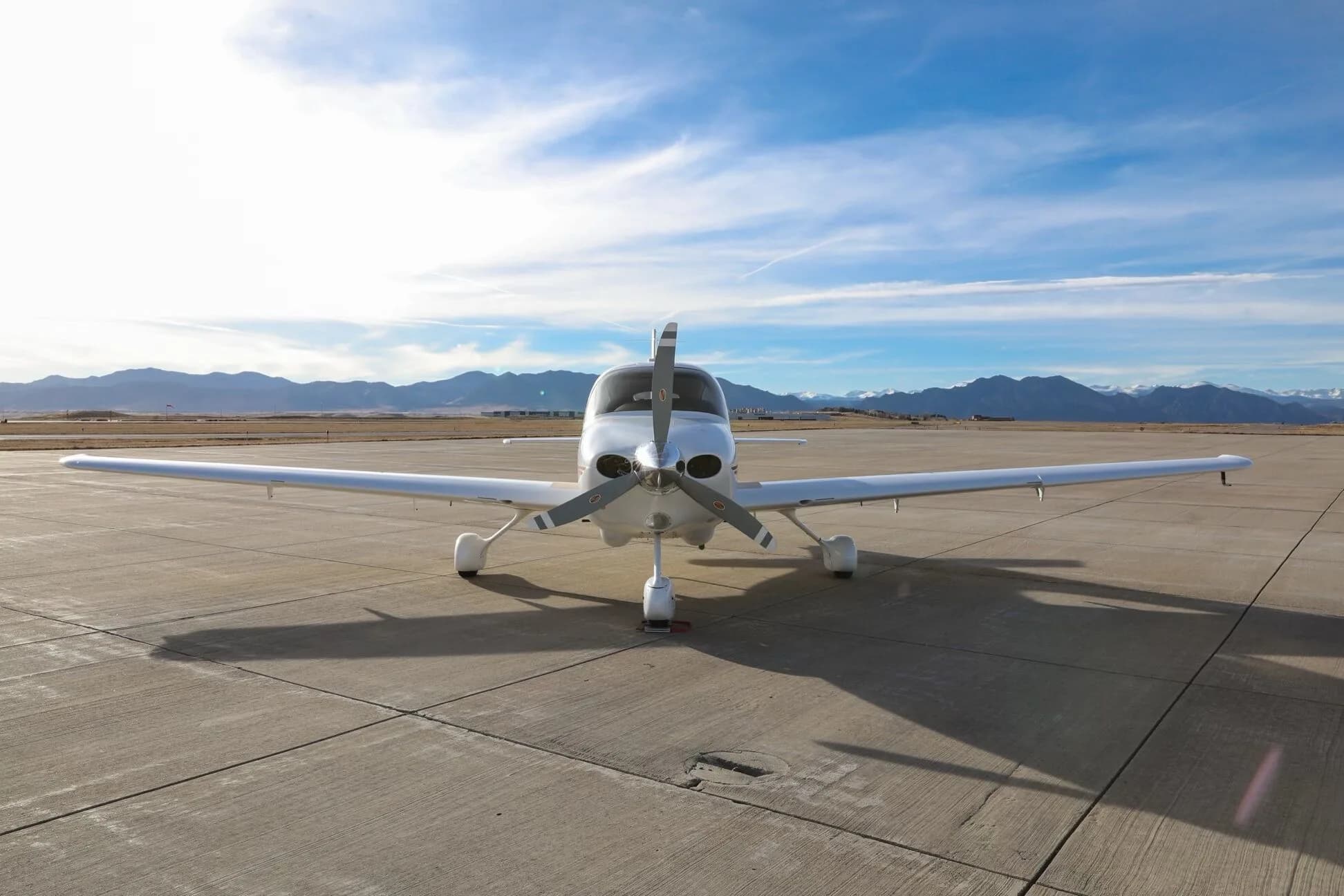 Front view of a white Cirrus SR20 aircraft with a three-bladed propeller, parked on an airfield with mountains in the distance.