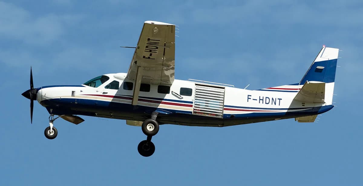 A white and blue Cessna Caravan is shown in flight against a blue sky.