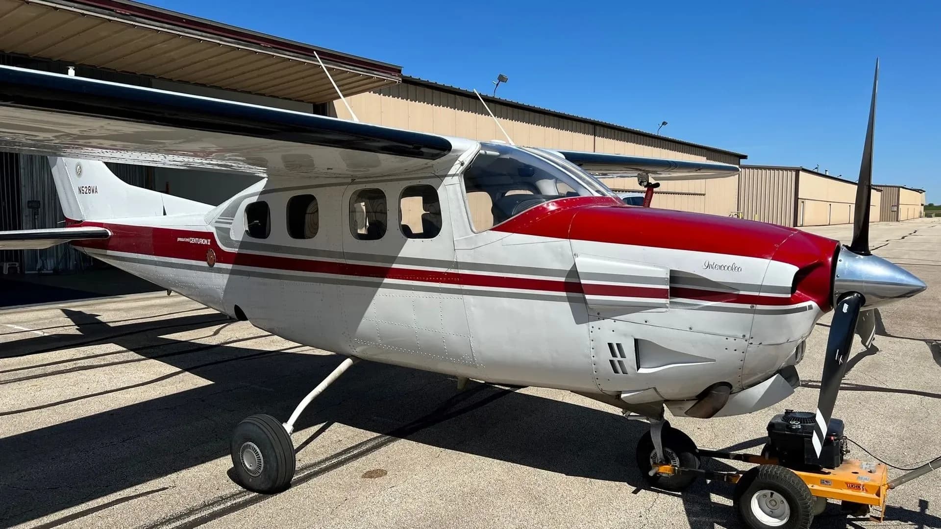 A white and red Cessna P210 Centurion II aircraft with tail number N528WA is parked on an airfield tarmac with hangars in the background.