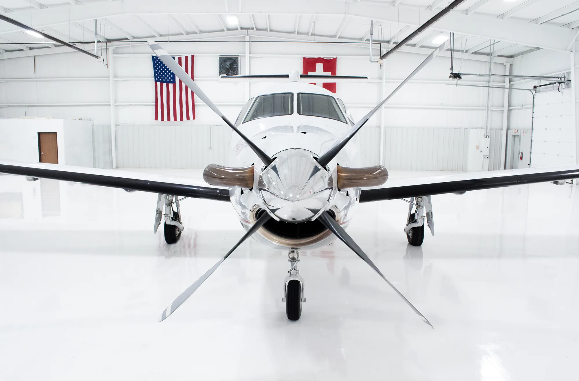Front view of a white Pilatus PC-12 aircraft with a large propeller, parked in a hangar with flags in the background.