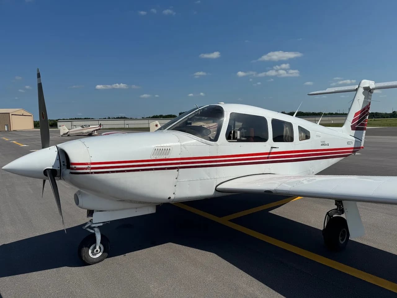 A white Piper Arrow II aircraft with red and brown stripes parked on an asphalt tarmac, viewed from the front left.
