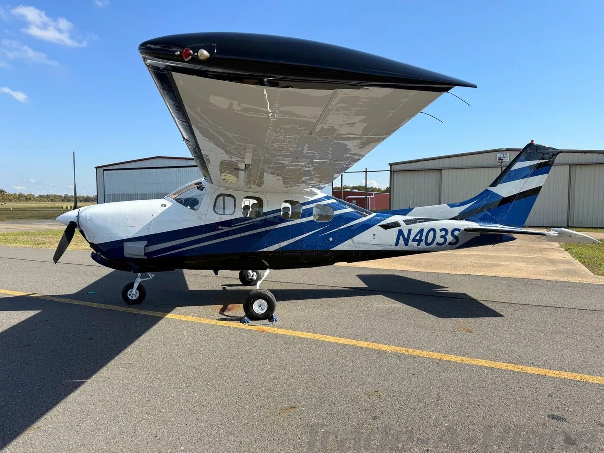 A white and blue Cessna 210 Centurion aircraft (N403S) parked on an asphalt ramp with hangars in the background.