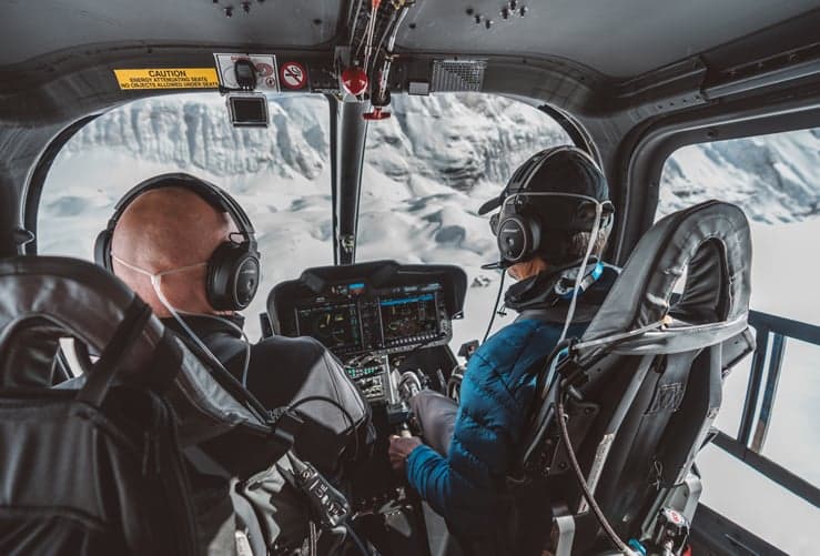 View from the back seat of a helicopter cockpit, showing the pilots and instrument panel.