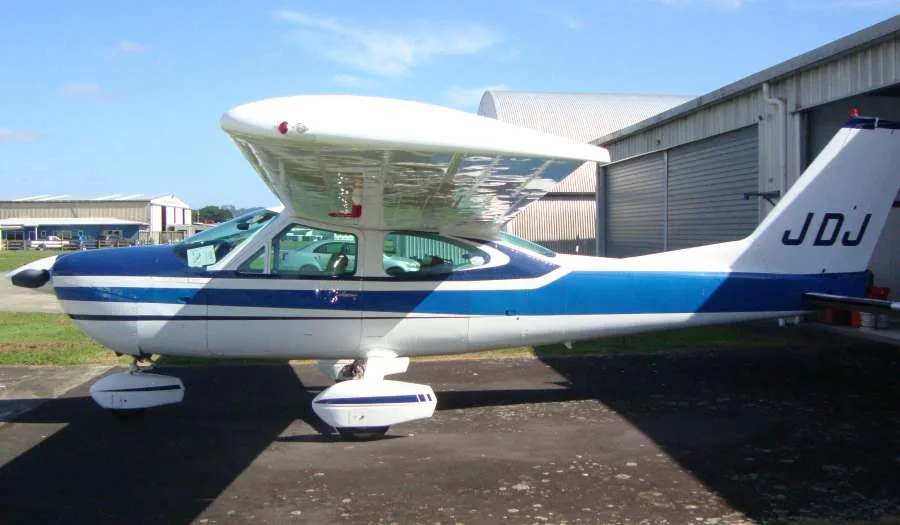 A white and blue single-engine propeller aircraft, likely a Cessna, with tail number JDJ, parked on an airfield next to a hangar.