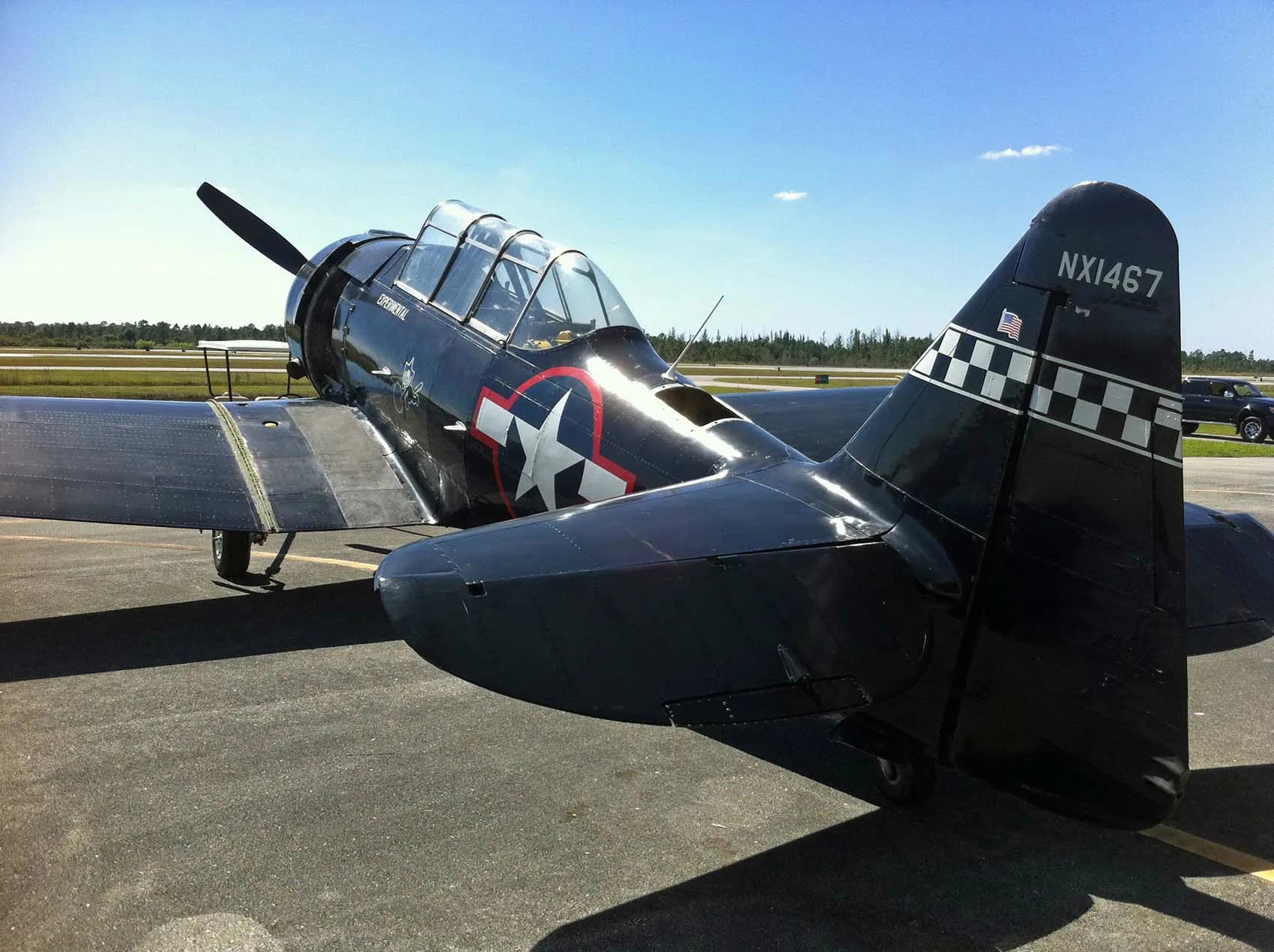 A rear-quarter view of a black warbird aircraft with US star insignia and registration NX1467, parked on an airfield under a clear sky.