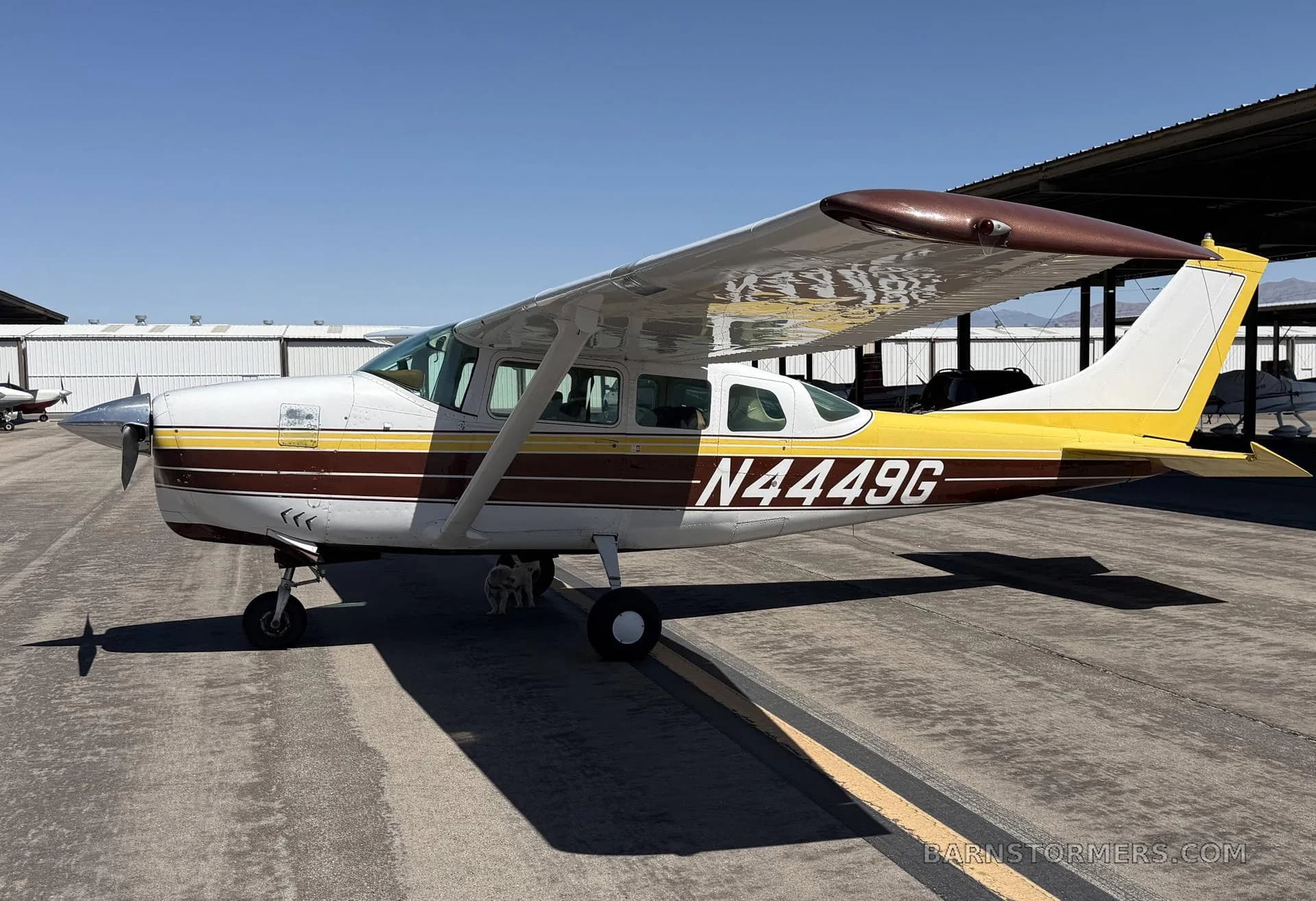 A white, yellow, and brown Cessna 182 aircraft with registration N4449G is parked on an asphalt apron under a clear blue sky.